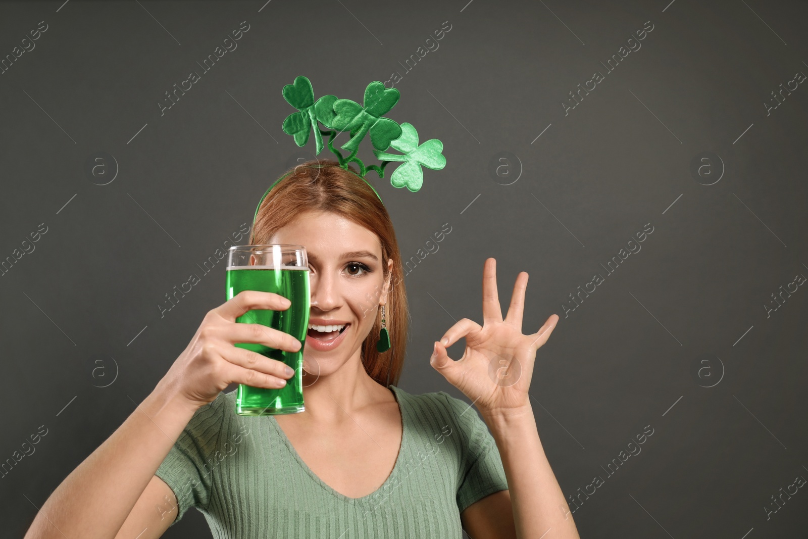 Photo of Young woman with clover headband and green beer on grey background. St. Patrick's Day celebration