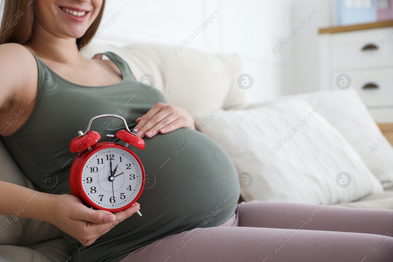 Photo of Young pregnant woman holding alarm clock near her belly at home, closeup. Time to give birth