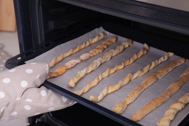 Photo of Woman taking baking sheet with homemade breadsticks out of oven, closeup. Cooking traditional grissini