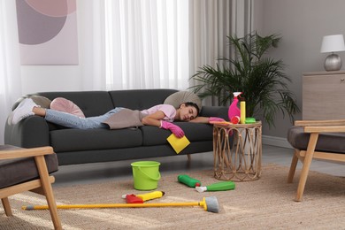 Tired young woman sleeping on sofa and cleaning supplies in living room