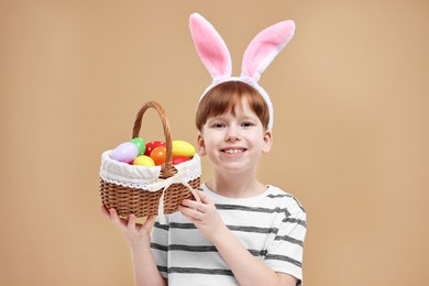 Easter celebration. Cute little boy with bunny ears and wicker basket full of painted eggs on dark beige background