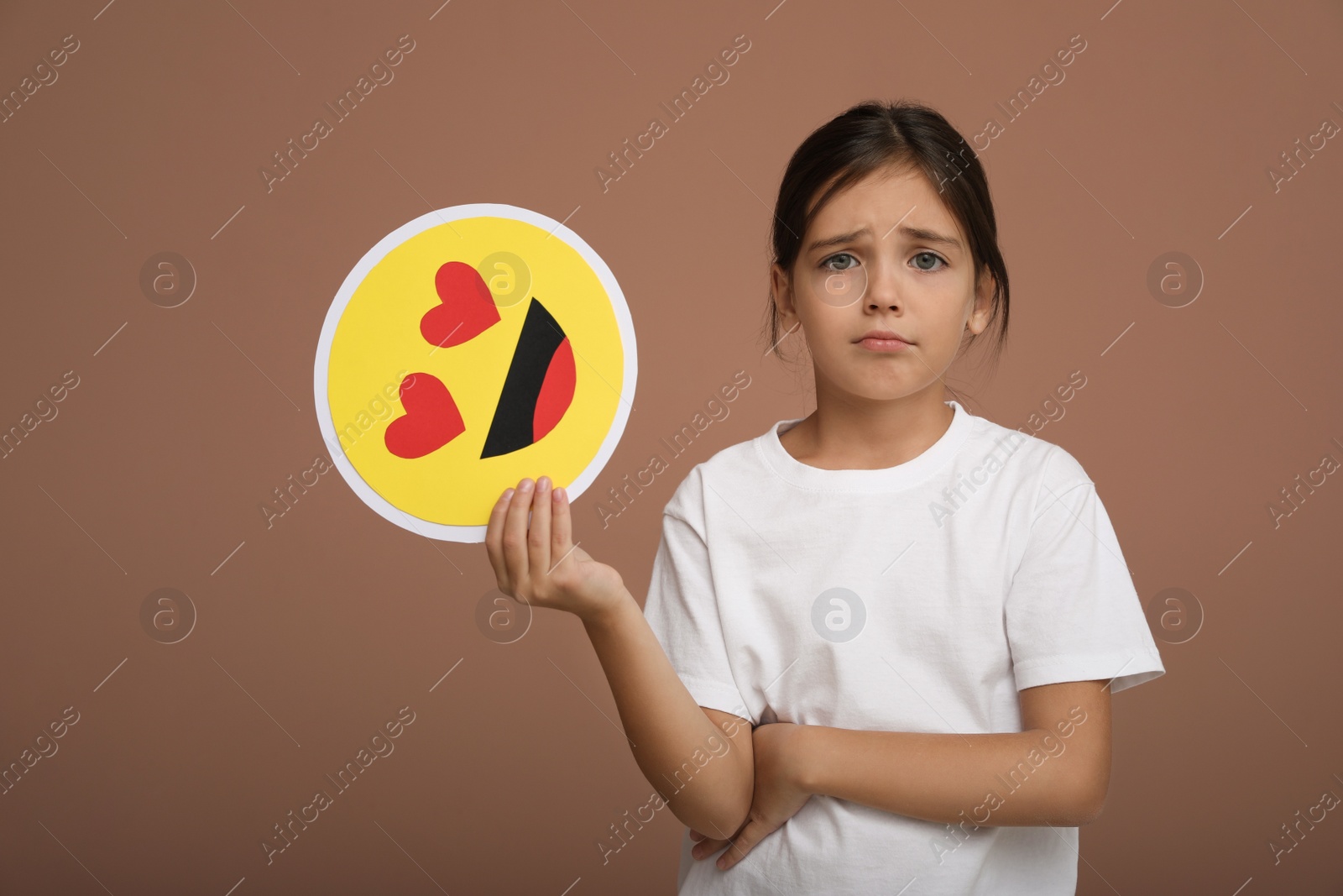 Photo of Sad little girl holding face with heart eyes emoji on pale pink background