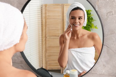Photo of Young woman cleaning her face with cotton pad near mirror in bathroom