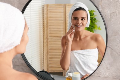 Young woman cleaning her face with cotton pad near mirror in bathroom