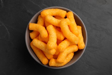 Photo of Many tasty cheesy corn puffs in bowl on black table, top view