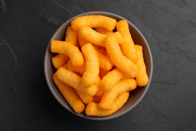 Many tasty cheesy corn puffs in bowl on black table, top view