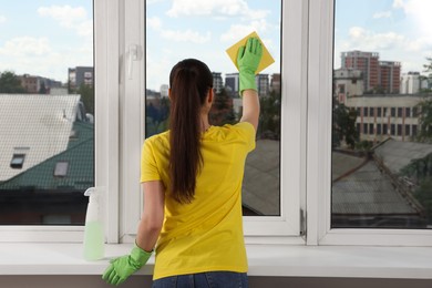 Photo of Woman cleaning window glass with sponge cloth indoors, back view