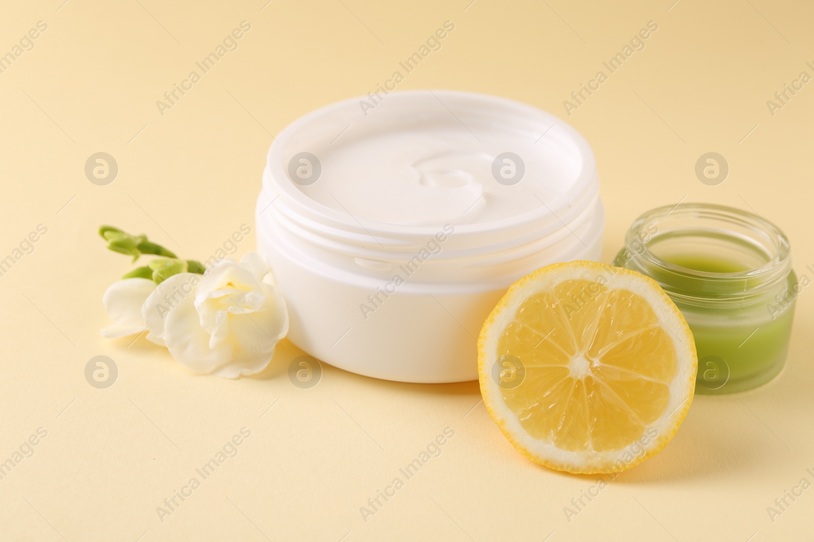 Photo of Moisturizing cream in open jars, lemon and freesia flower on beige background. Body care products