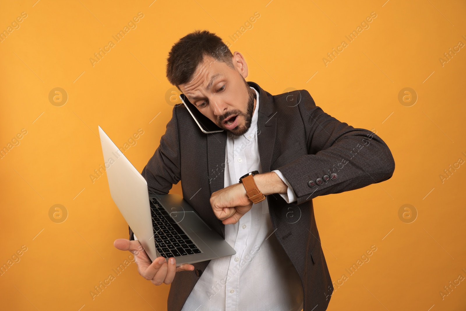 Photo of Emotional man with laptop checking time while talking on phone against orange background. Being late concept