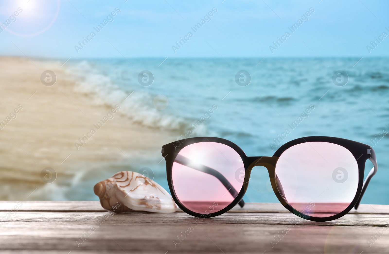 Image of Shell and stylish sunglasses on wooden table near sea with sandy beach