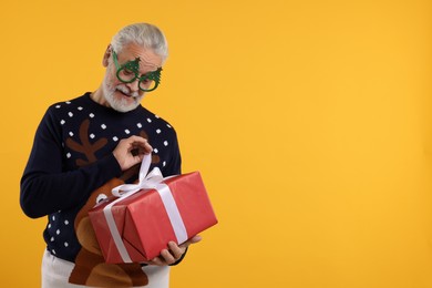 Photo of Senior man in Christmas sweater and funny glasses opening gift against orange background. Space for text