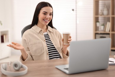 Photo of Young woman with cup of coffee using video chat during webinar at table in room