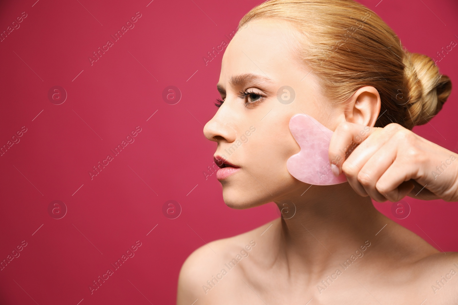 Photo of Beautiful young woman doing facial massage with gua sha tool on pink background, closeup. Space for text