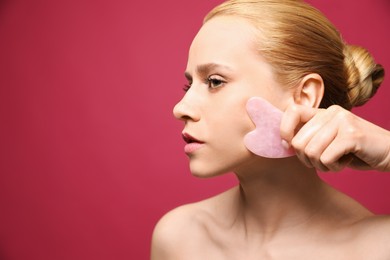 Photo of Beautiful young woman doing facial massage with gua sha tool on pink background, closeup. Space for text