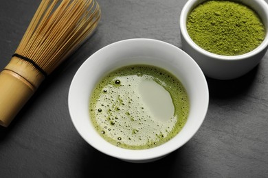 Photo of Cup of fresh matcha tea, green powder and bamboo whisk on black table, closeup