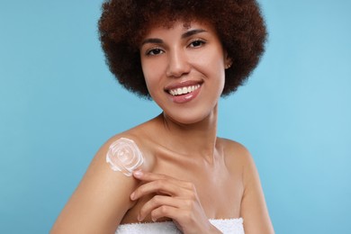 Beautiful young woman applying body cream onto shoulder on light blue background