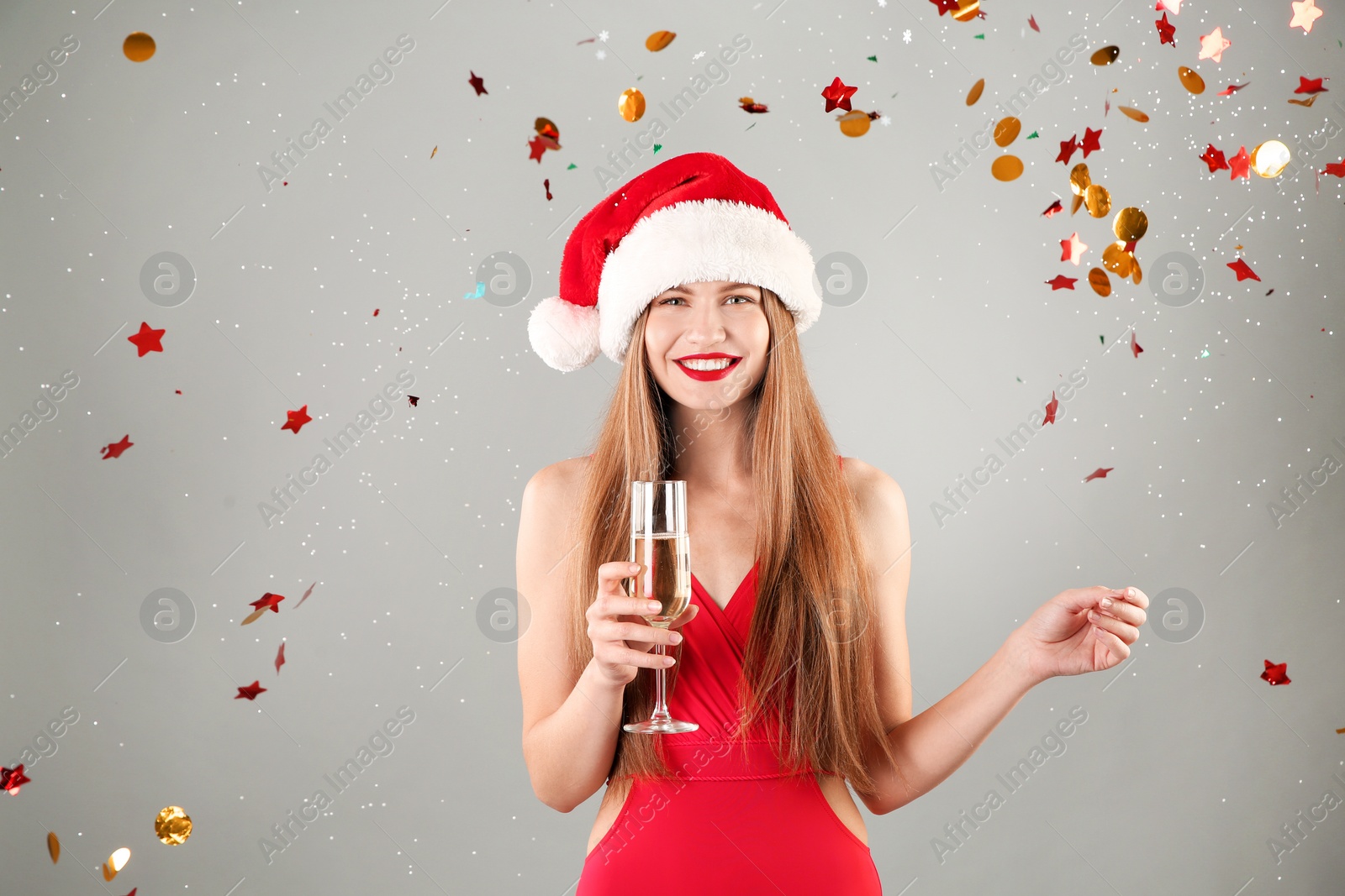 Photo of Young beautiful woman in Santa hat and swimsuit holding glass of champagne on grey background. Christmas celebration