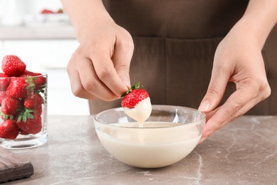 Photo of Woman dipping ripe strawberry into bowl with white melted chocolate on table