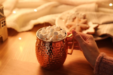 Photo of Woman holding cup of hot drink with marshmallows at wooden table, closeup. Magic Christmas atmosphere