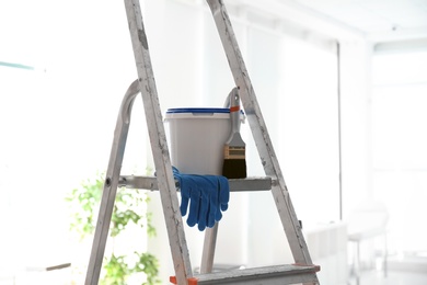 Photo of Bucket with paint, brush and gloves on step ladder in light room