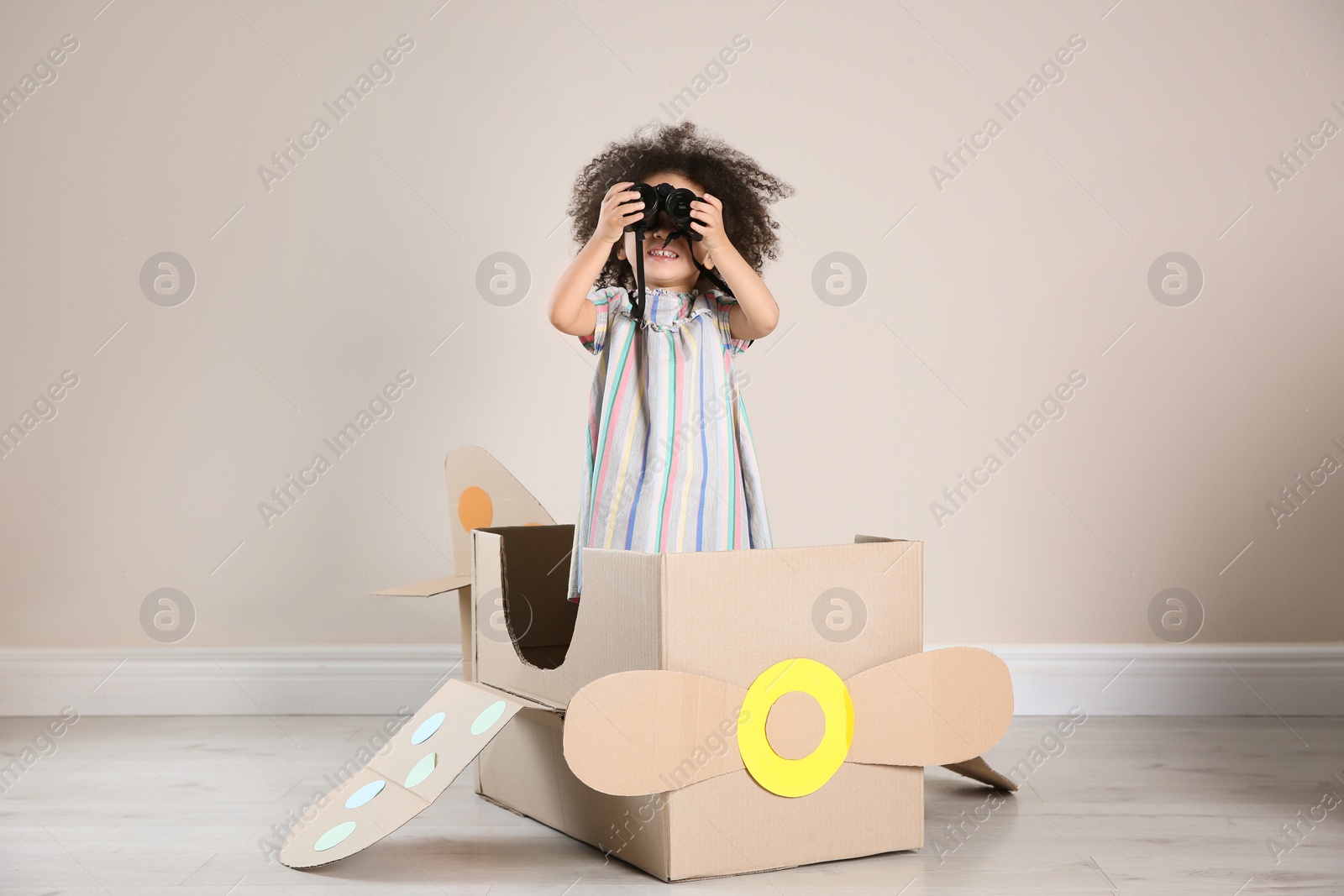 Photo of Cute African American child playing with cardboard plane and binoculars near beige wall