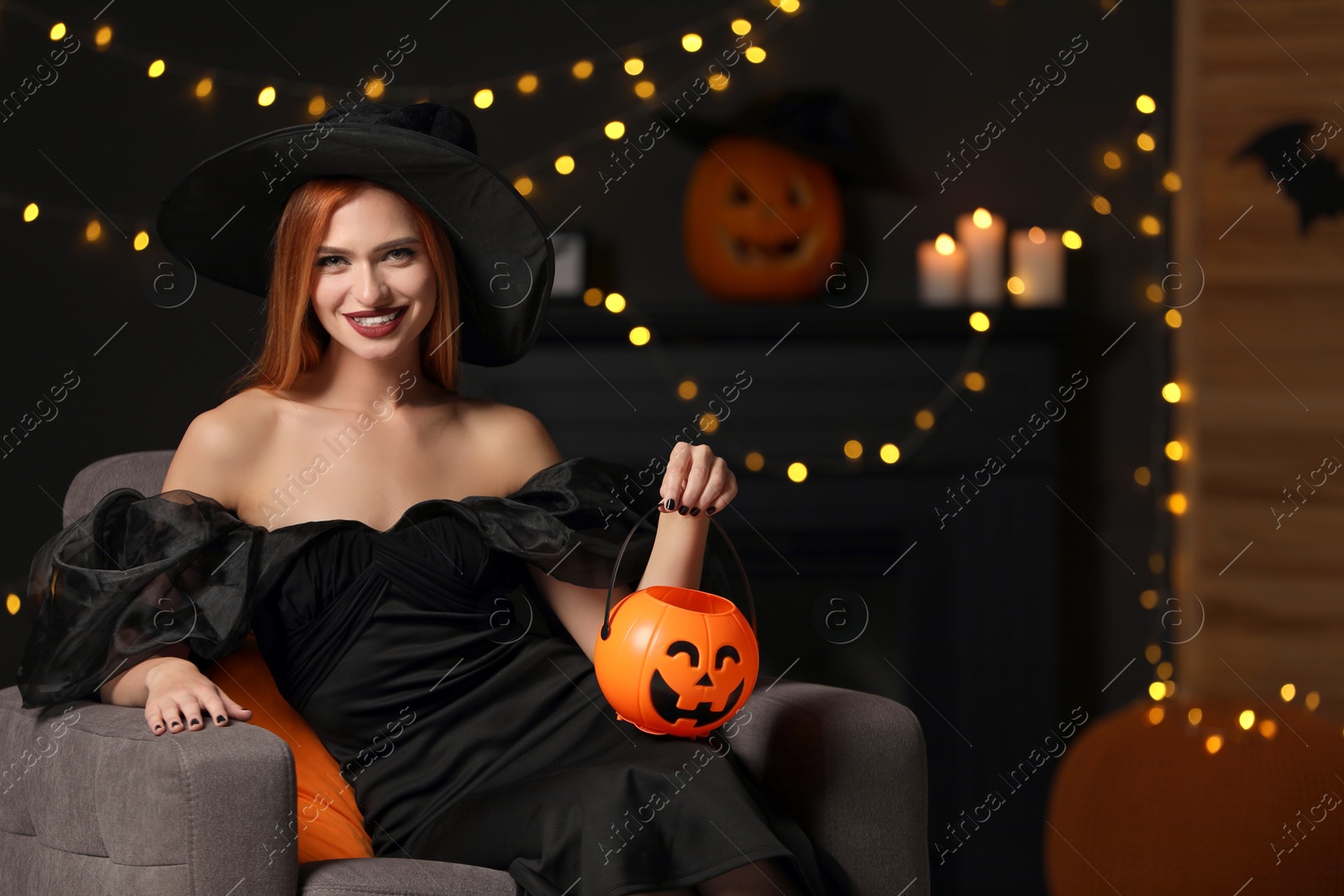 Photo of Young woman in scary witch costume with pumpkin bucket against blurred lights indoors, space for text. Halloween celebration