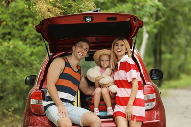 Happy family sitting in car trunk with suitcases near forest