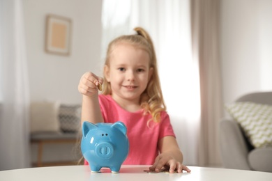 Photo of Cute girl putting coin into piggy bank at table in living room. Saving money