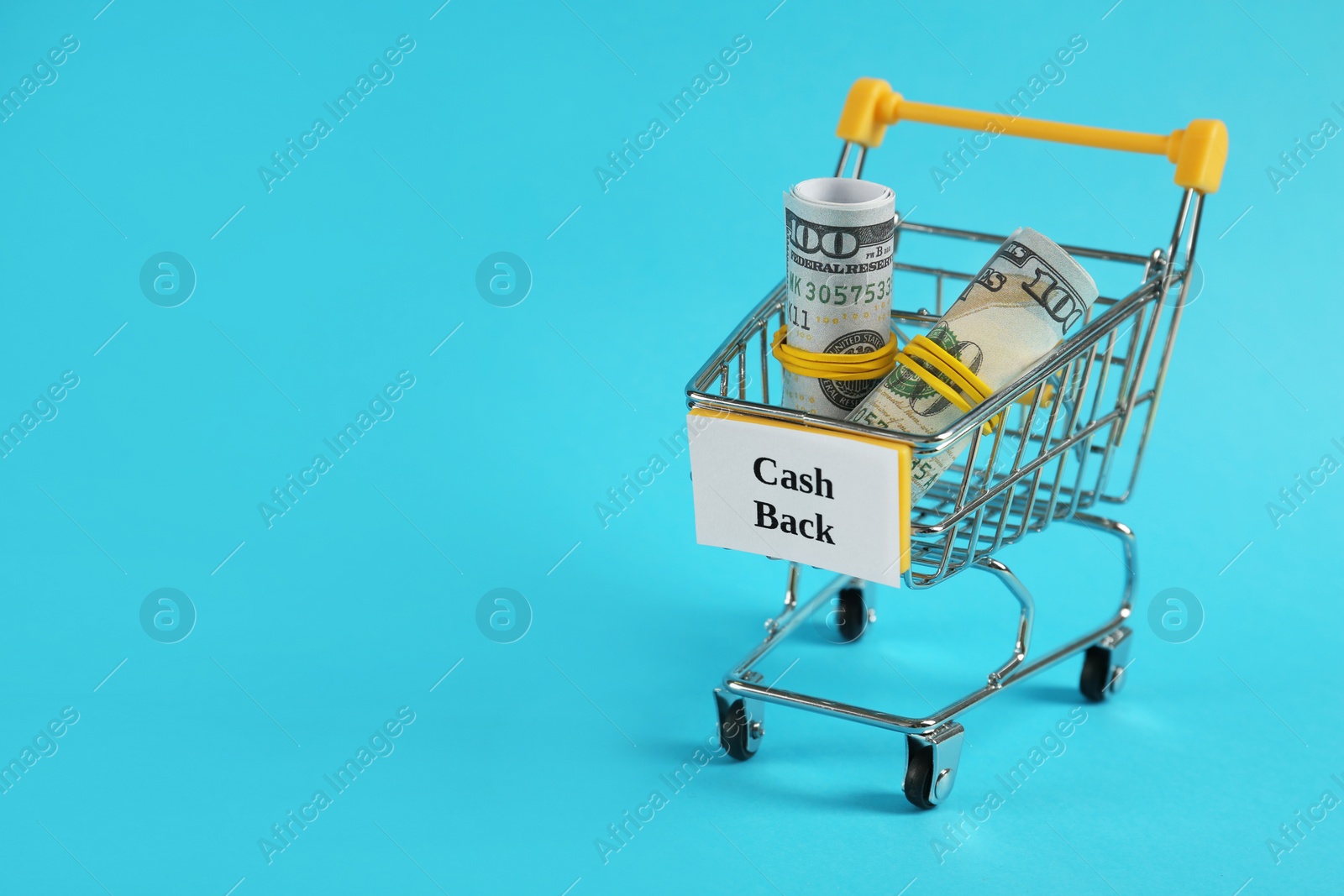 Photo of Rolled dollar banknotes in shopping cart on light blue background, space for text. Cashback concept