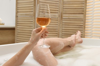 Photo of Woman with glass of wine taking bath in tub indoors, closeup