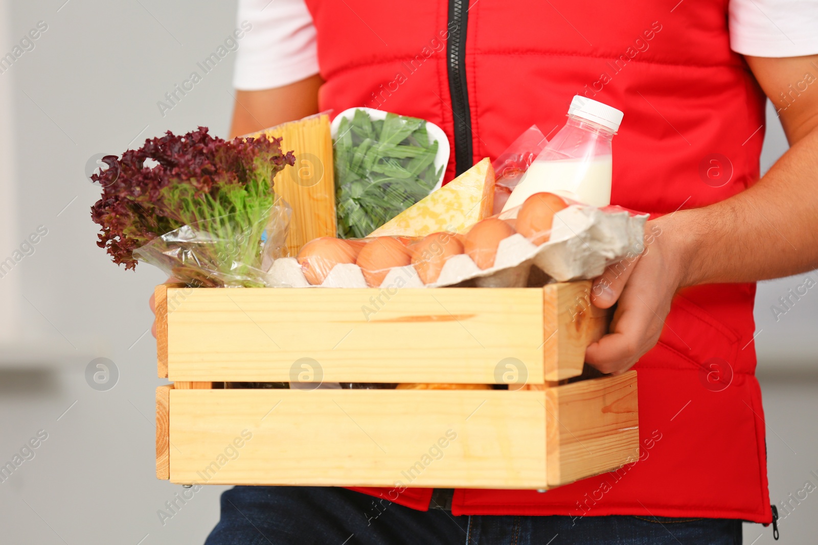 Photo of Male courier holding wooden crate with products on light background, closeup. Food delivery service