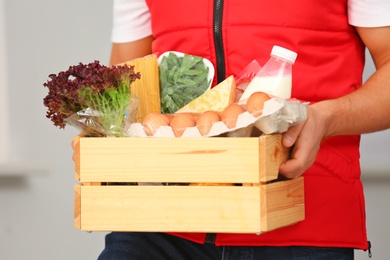 Male courier holding wooden crate with products on light background, closeup. Food delivery service