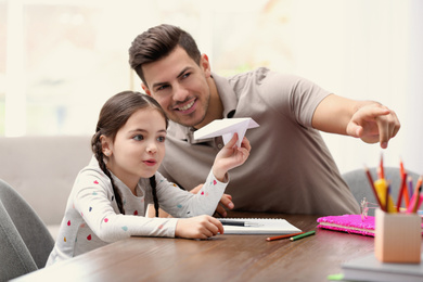 Photo of Father and daughter playing with paper plane while doing homework together at table indoors