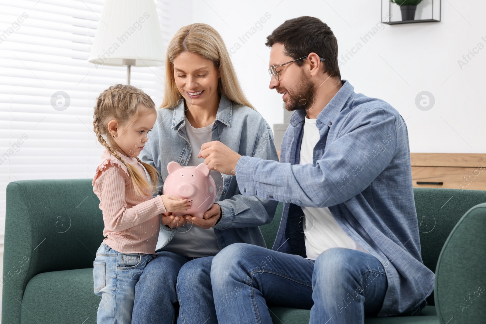 Photo of Planning budget together. Little girl with her parents putting coin into piggybank at home