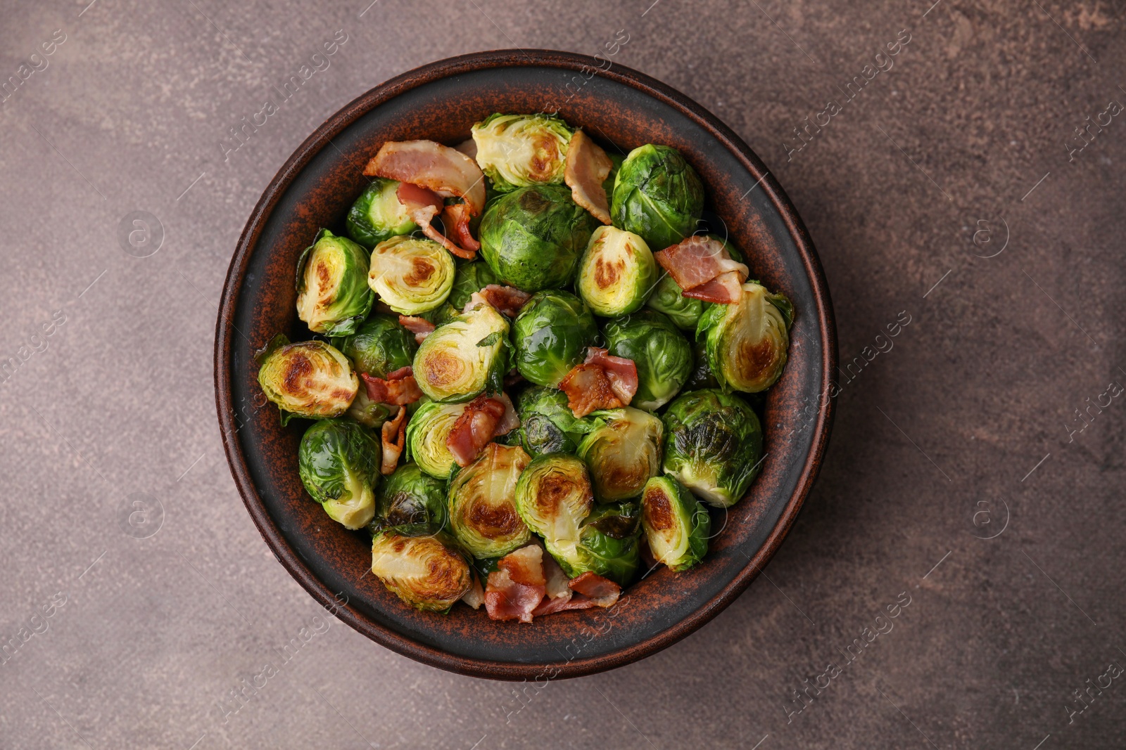 Photo of Delicious roasted Brussels sprouts and bacon in bowl on brown table, top view