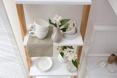 Photo of Beautiful jasmine flowers and tableware on shelving unit in kitchen, above view