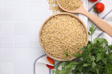 Photo of Raw bulgur in bowl, spoon, vegetables and parsley on white tiled table, flat lay. Space for text
