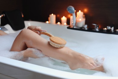 Photo of Woman rubbing leg with brush while taking bubble bath, closeup. Romantic atmosphere