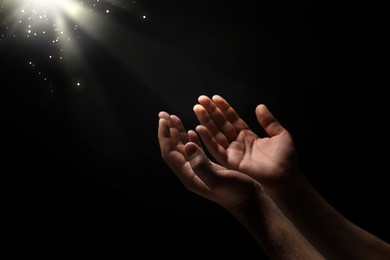 Christian man stretching hands towards holy light in darkness, closeup. Prayer and belief