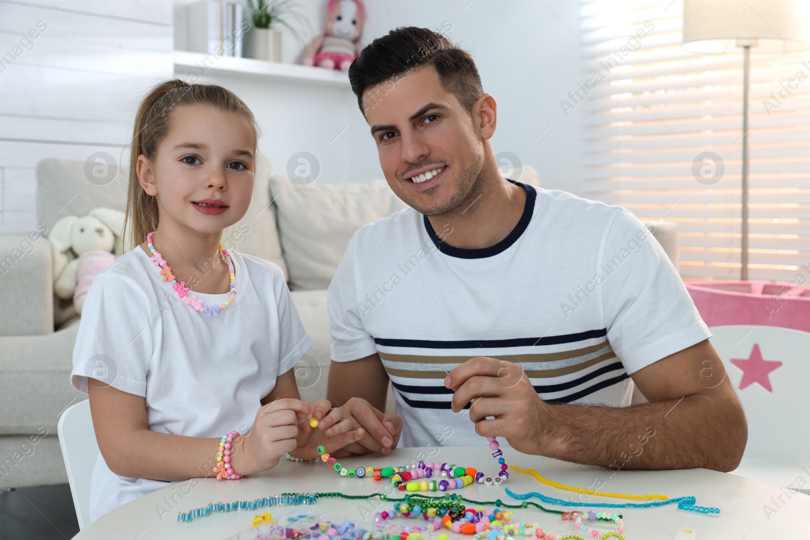 Photo of Happy father with his cute daughter making beaded jewelry at table in room