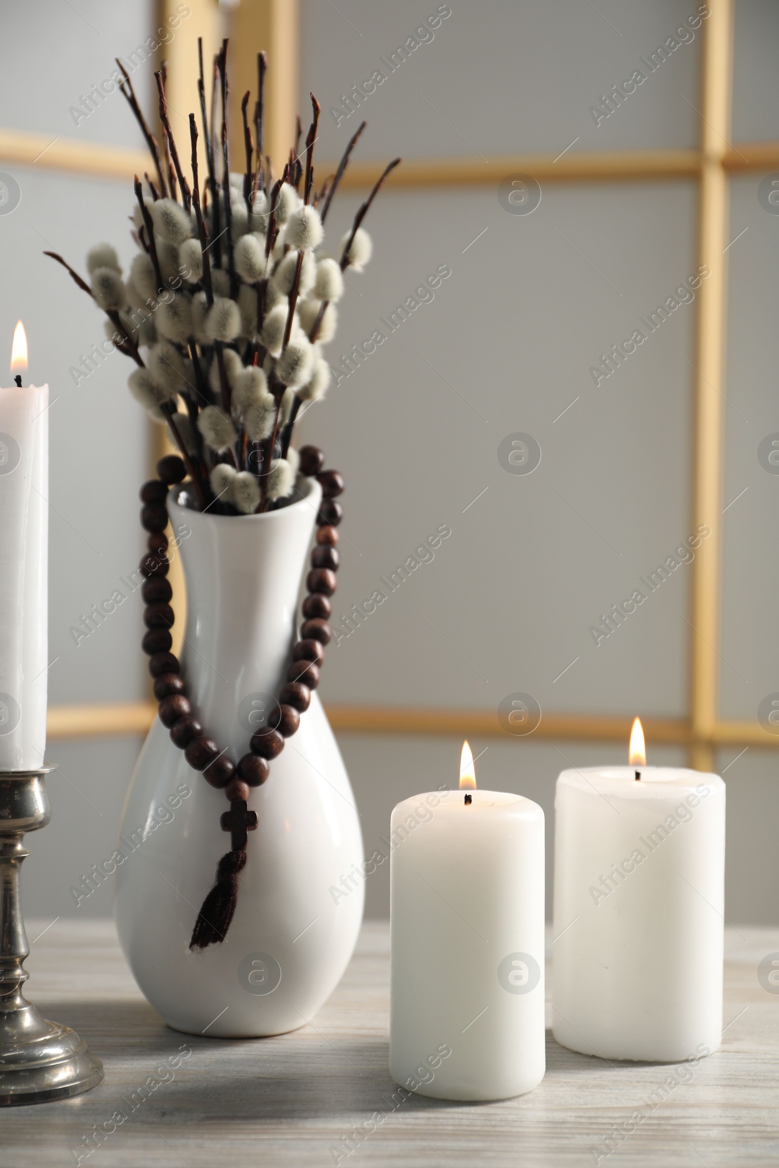 Photo of Rosary beads, burning candles and vase of willow branches on wooden table
