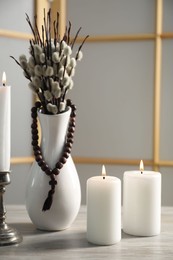 Rosary beads, burning candles and vase of willow branches on wooden table