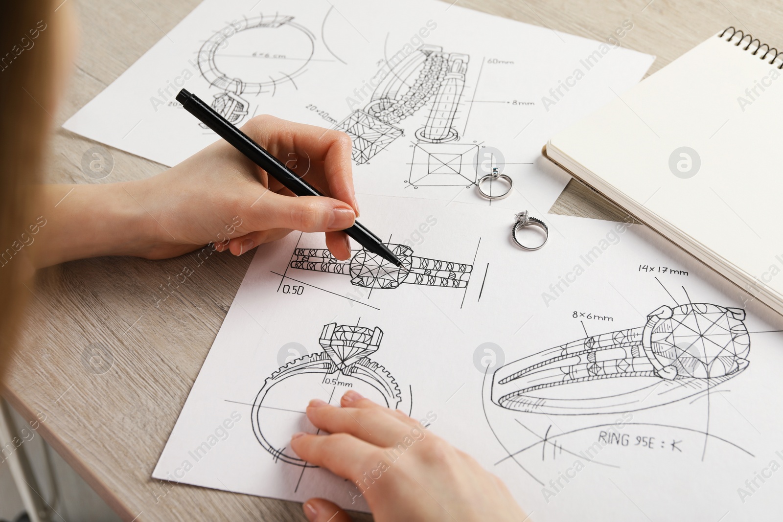 Photo of Jeweler drawing sketch of elegant ring on paper at wooden table, closeup