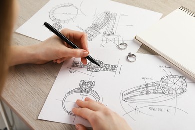Photo of Jeweler drawing sketch of elegant ring on paper at wooden table, closeup