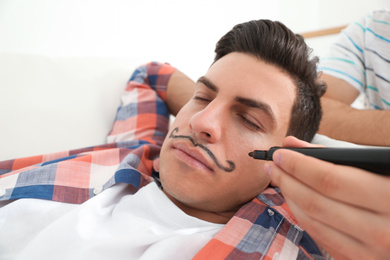 Young man drawing mustache on face of sleeping friend, closeup. April fool's day