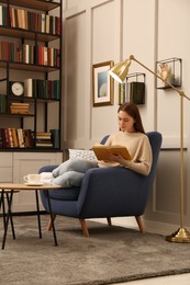 Young woman reading book in armchair indoors. Home library