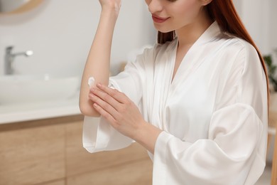Photo of Young woman applying body cream onto elbow in bathroom, closeup. Space for text