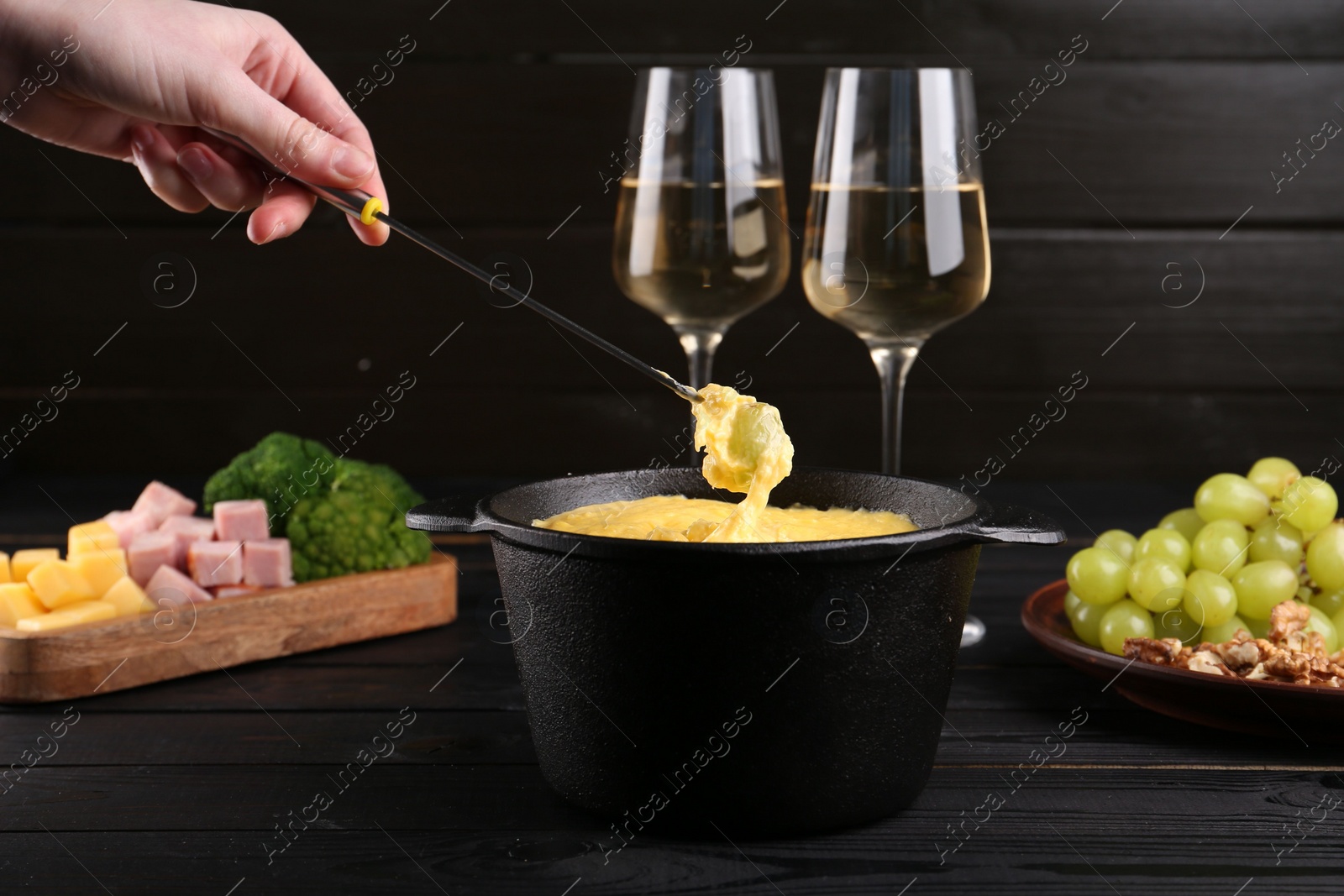 Photo of Woman dipping grape into fondue pot with melted cheese at black wooden table, closeup