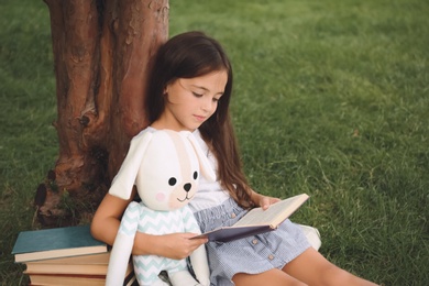Photo of Cute little girl with toy reading book on green grass near tree in park
