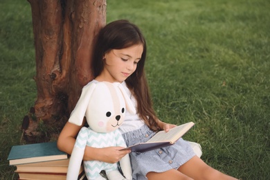 Cute little girl with toy reading book on green grass near tree in park